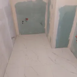 Łazienka w trakcie remontu, widoczne płytki podłogowe imitujące marmur, ściany z częściowo nałożonym tynkiem i wyprowadzonymi instalacjami elektrycznymi.