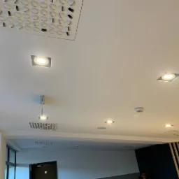 Biały sufit z różnymi typami oświetlenia, w tym kwadratowe lampy LED, kratka wentylacyjna i otwór rewizyjny z regularnym wzorem okręgów.