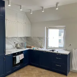 Nowoczesna kuchnia po remoncie: granatowe szafki z białymi górnymi szafkami, marmurowy backsplash, złota armatura, parkiet w jodełkę i biały grzejnik pod oknem.