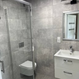 ASKI GROUP - Nowoczesna łazienka w szarościach: kabina prysznicowa ze szklanymi drzwiami, podwieszana toaleta, biała szafka z umywalką i lustro z oświetleniem.