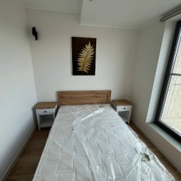 ASKI GROUP - Jasna sypialnia po remoncie: drewniane łóżko z materacem w folii, szafki nocne, obraz z motywem liścia i duże okno z widokiem na kamienicę.