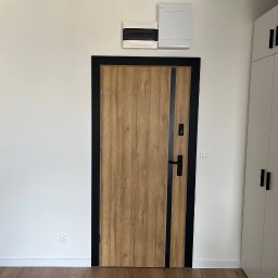 ASKI GROUP - Nowoczesne wnętrze: drewniane drzwi z czarną ramą, białe ściany i szafa w zabudowie. Minimalistyczny design, jasne kolory, widoczna skrzynka elektryczna.