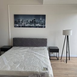 ASKI GROUP - Jasne wnętrze sypialni po remoncie: białe ściany z dekoracyjną listwą, łóżko z szarym zagłówkiem, nocne stoliki i lampa. Na ścianie obraz z panoramą miasta.