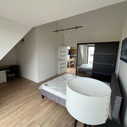 ASKI GROUP - Jasne wnętrze pokoju po odświeżeniu ścian: drewniana podłoga, białe ściany, biurko pod skosem, łóżko z lampą i szafa z lustrem. Widok na kuchnię w tle.