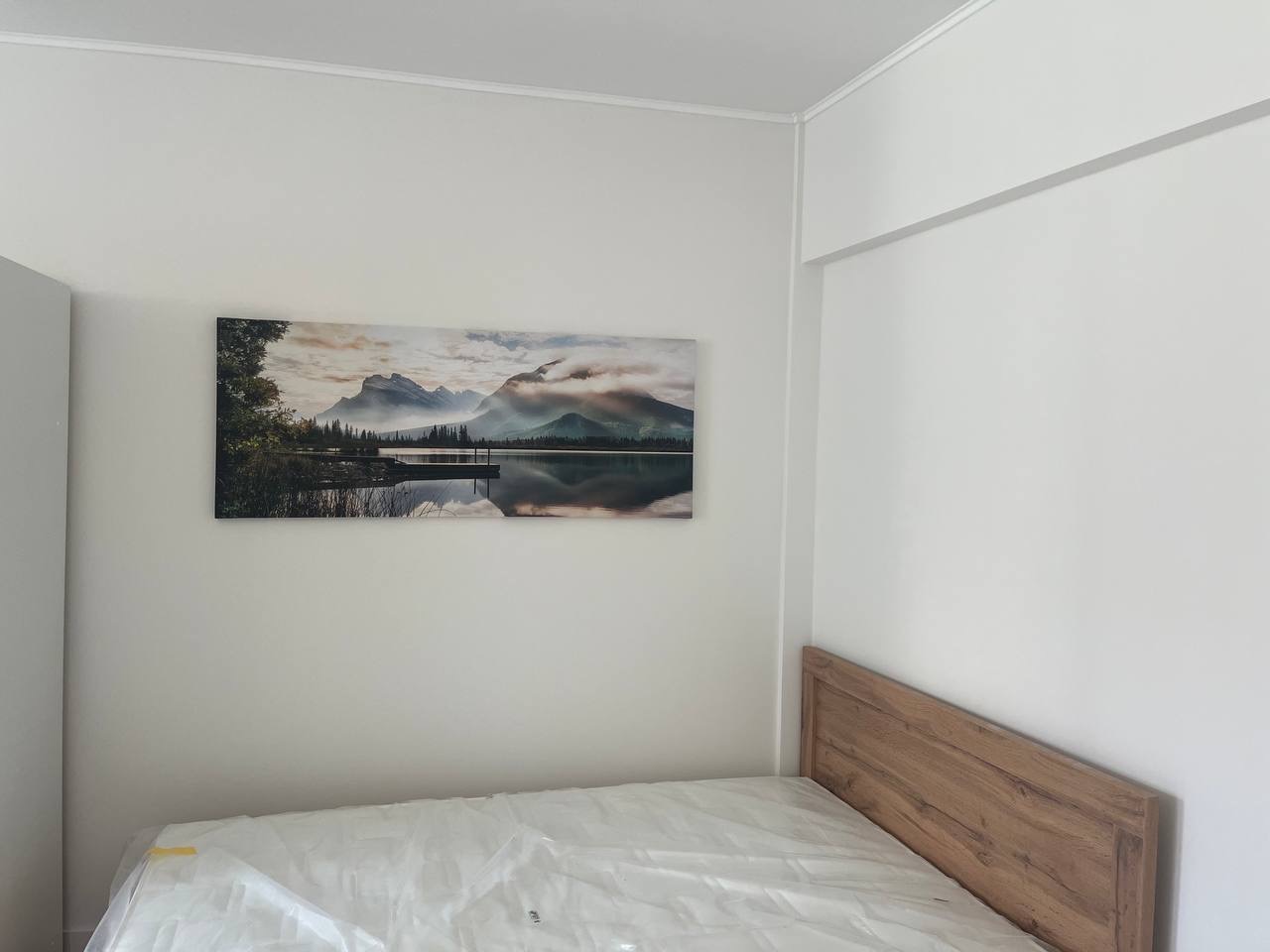 Biała ściana z obrazem górskiego krajobrazu nad łóżkiem z drewnianym wezgłowiem. Minimalistyczne wnętrze sypialni po remoncie, z listwą przysufitową.