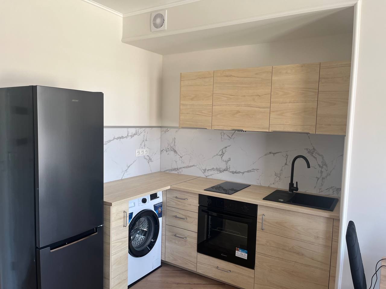 Jasna kuchnia po remoncie: drewnopodobne szafki, marmurowy backsplash, czarny zlew i sprzęt AGD. Pralka w zabudowie obok lodówki.