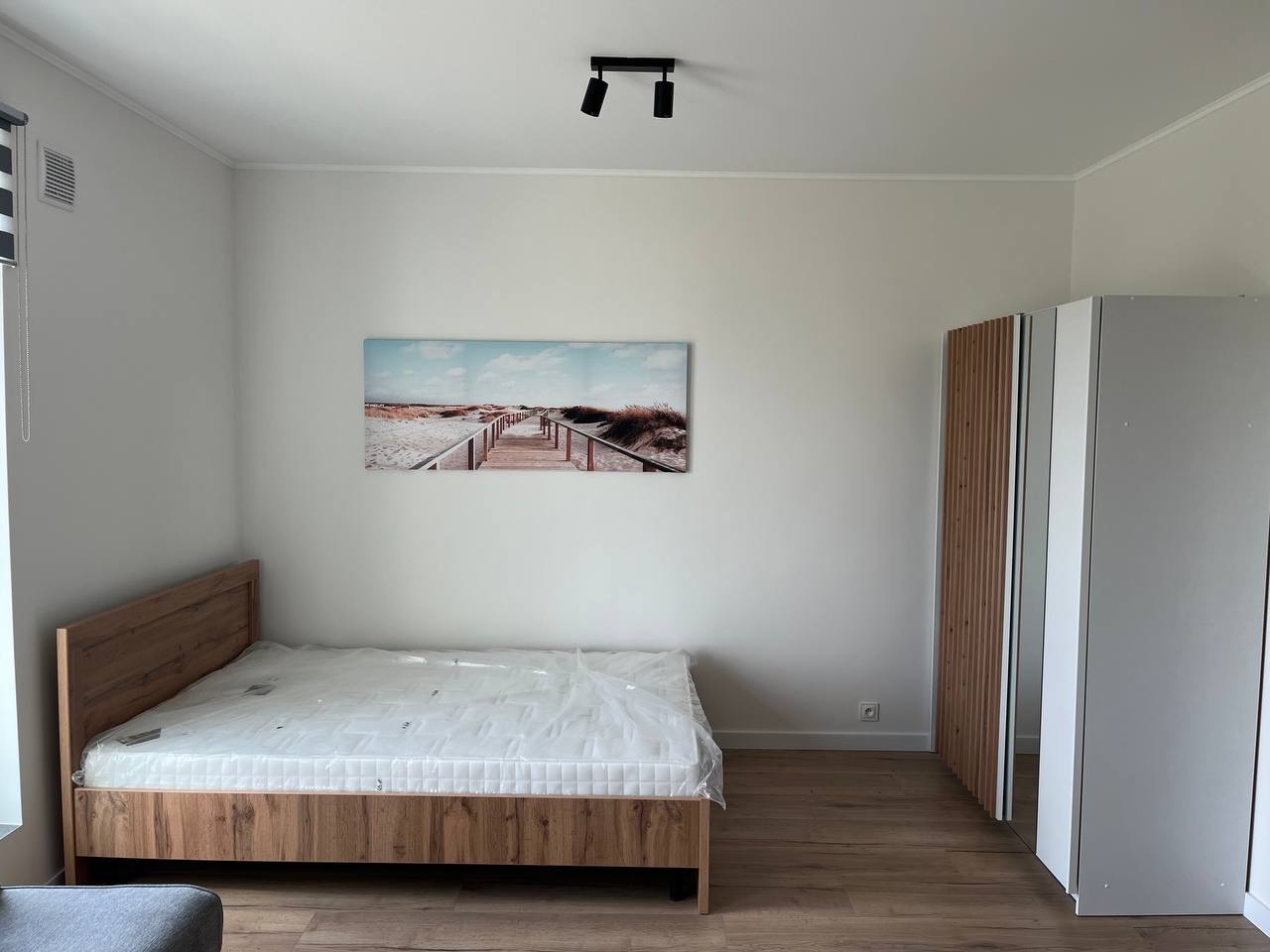 Jasne wnętrze sypialni po remoncie: drewniane łóżko z materacem w folii, szafa z drewnianymi elementami i obraz z motywem plaży na białej ścianie. Minimalistyczny design.