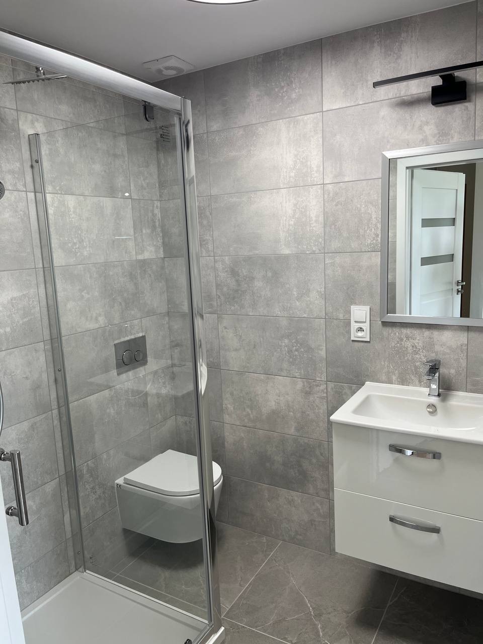 Nowoczesna łazienka w szarościach: kabina prysznicowa ze szklanymi drzwiami, podwieszana toaleta, biała szafka z umywalką i lustro z oświetleniem.
