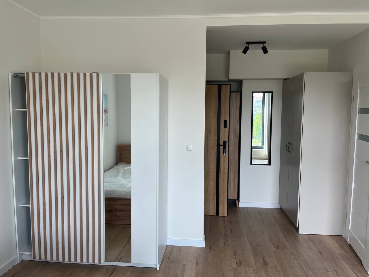 Jasne wnętrze z drewnianą podłogą i białymi meblami. Widoczna szafa z lustrem i drewnianymi lamelami, drzwi z elementami drewna oraz oświetlenie sufitowe.