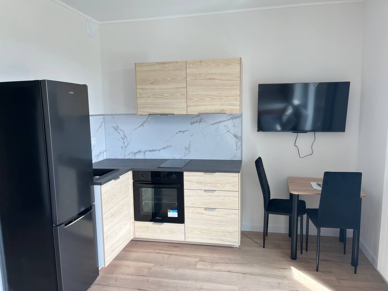 Nowoczesna, jasna kuchnia z drewnianymi szafkami, czarnym blatem i lodówką. Marmurowy panel nad blatem. Stół z krzesłami i telewizor na ścianie.
