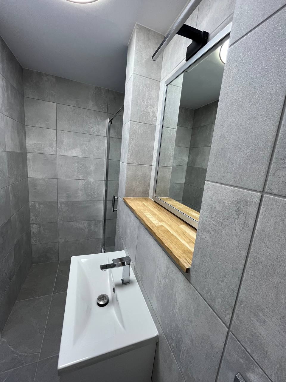 Szara łazienka z kabiną prysznicową, umywalką i lustrem z drewnianą półką. Minimalistyczny design, płytki imitujące beton, chromowana armatura. Nowoczesne wykończenie.