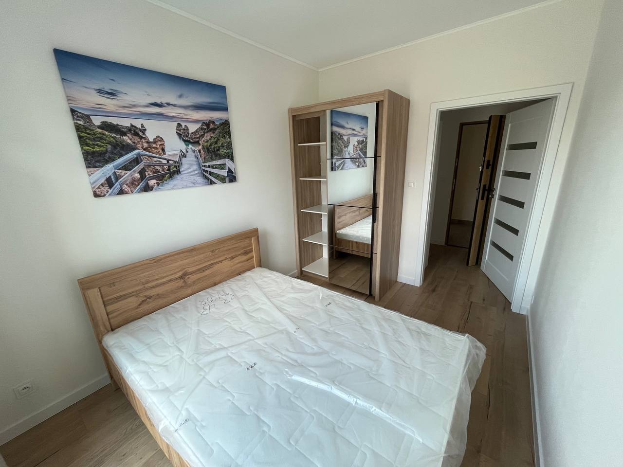 Jasne wnętrze pokoju z drewnianym łóżkiem, szafą z lustrem i obrazem nad wezgłowiem. Podłoga z paneli, białe ściany i drzwi w tle.