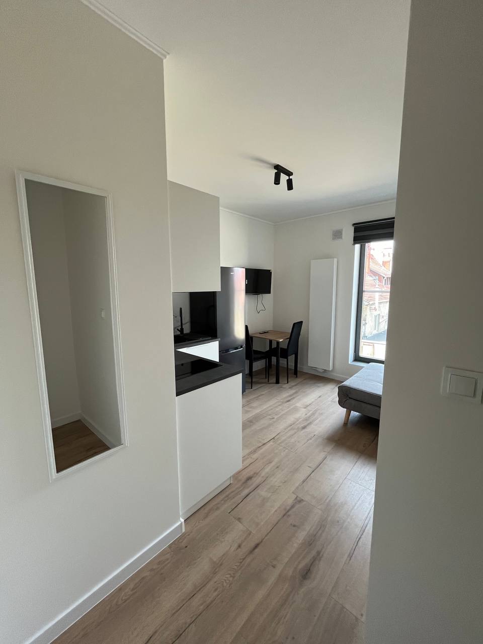 Jasne wnętrze kawalerki po remoncie: białe ściany, drewnopodobna podłoga, aneks kuchenny z czarnym blatem, stół z krzesłami, szara sofa przy oknie.