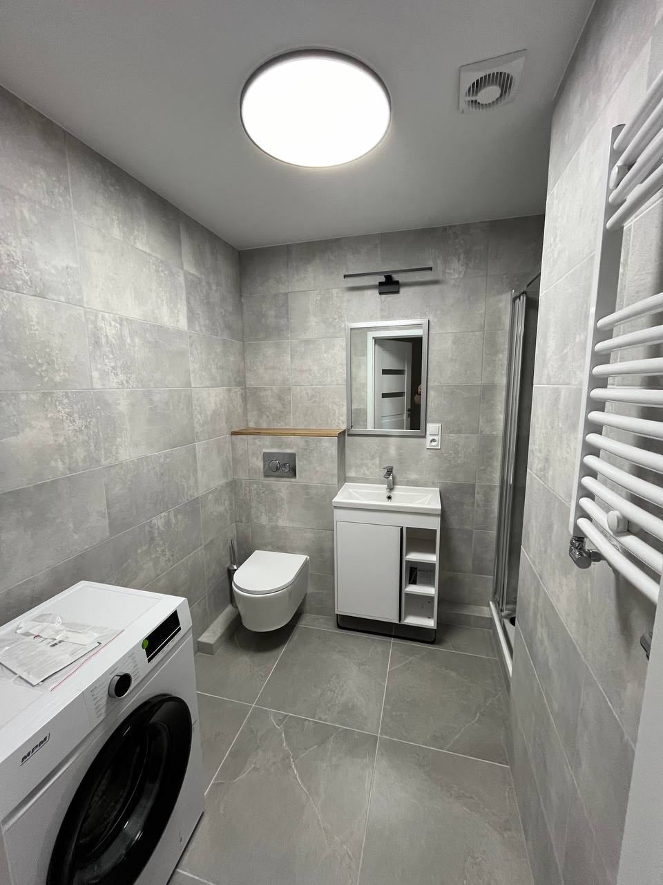 Szara łazienka po remoncie z pralką, toaletą, umywalką z szafką i grzejnikiem drabinkowym. Minimalistyczny design, płytki imitujące kamień, oświetlenie sufitowe.