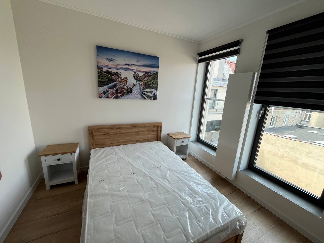 Jasna sypialnia z drewnianym łóżkiem i szafkami nocnymi. Na ścianie obraz z motywem morskim. Okna z roletami dzień-noc. Minimalistyczny wystrój wnętrza.