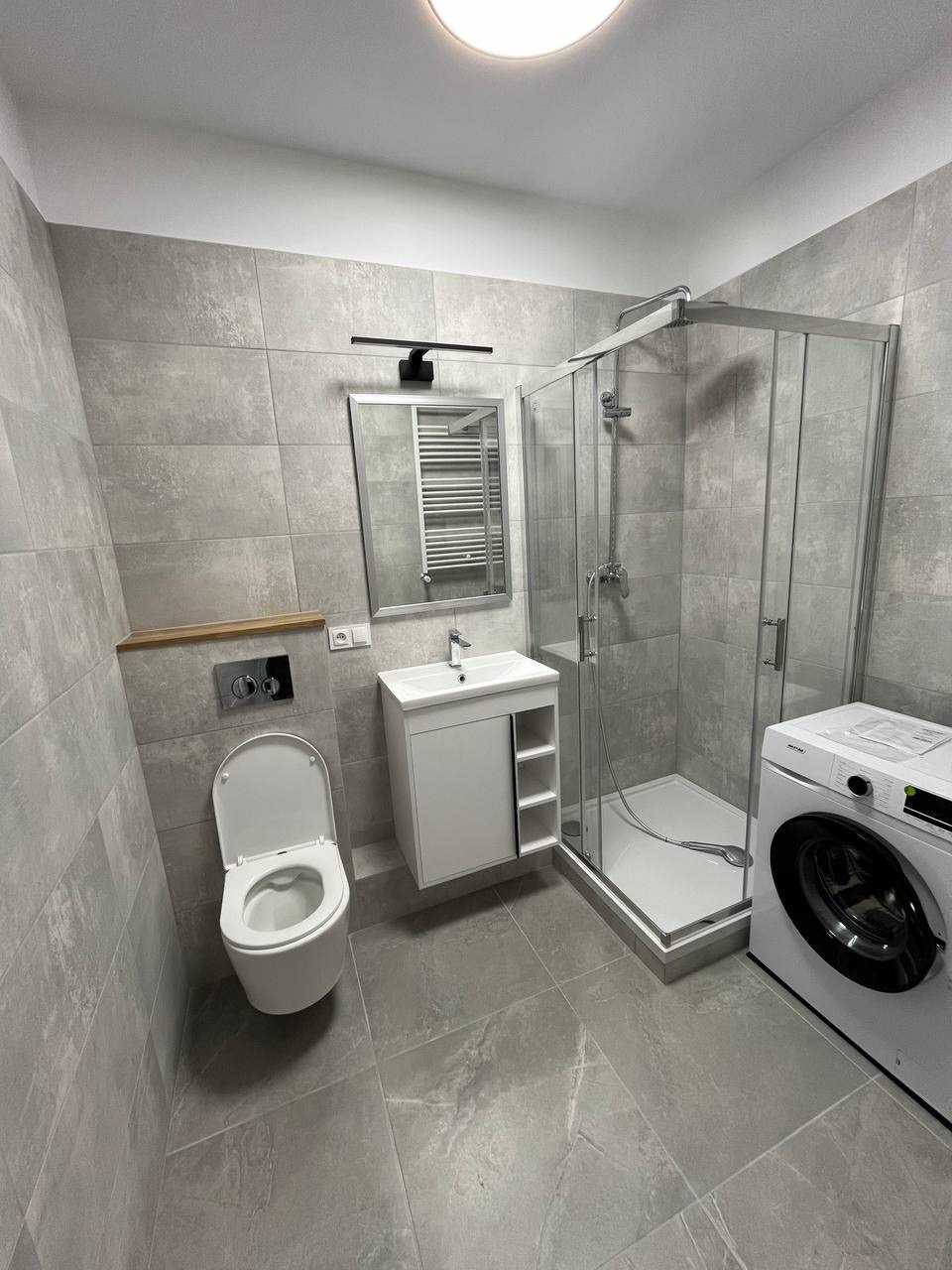 Wyremontowana łazienka w szarościach: kabina prysznicowa, umywalka z szafką, toaleta i pralka. Minimalistyczny design, płytki imitujące kamień, oświetlenie punktowe.