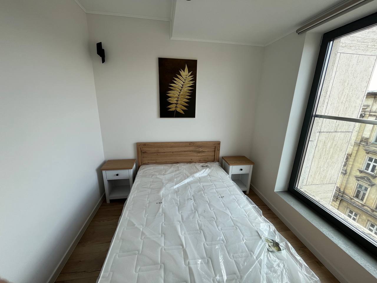 Jasna sypialnia po remoncie: drewniane łóżko z materacem w folii, szafki nocne, obraz z motywem liścia i duże okno z widokiem na kamienicę.