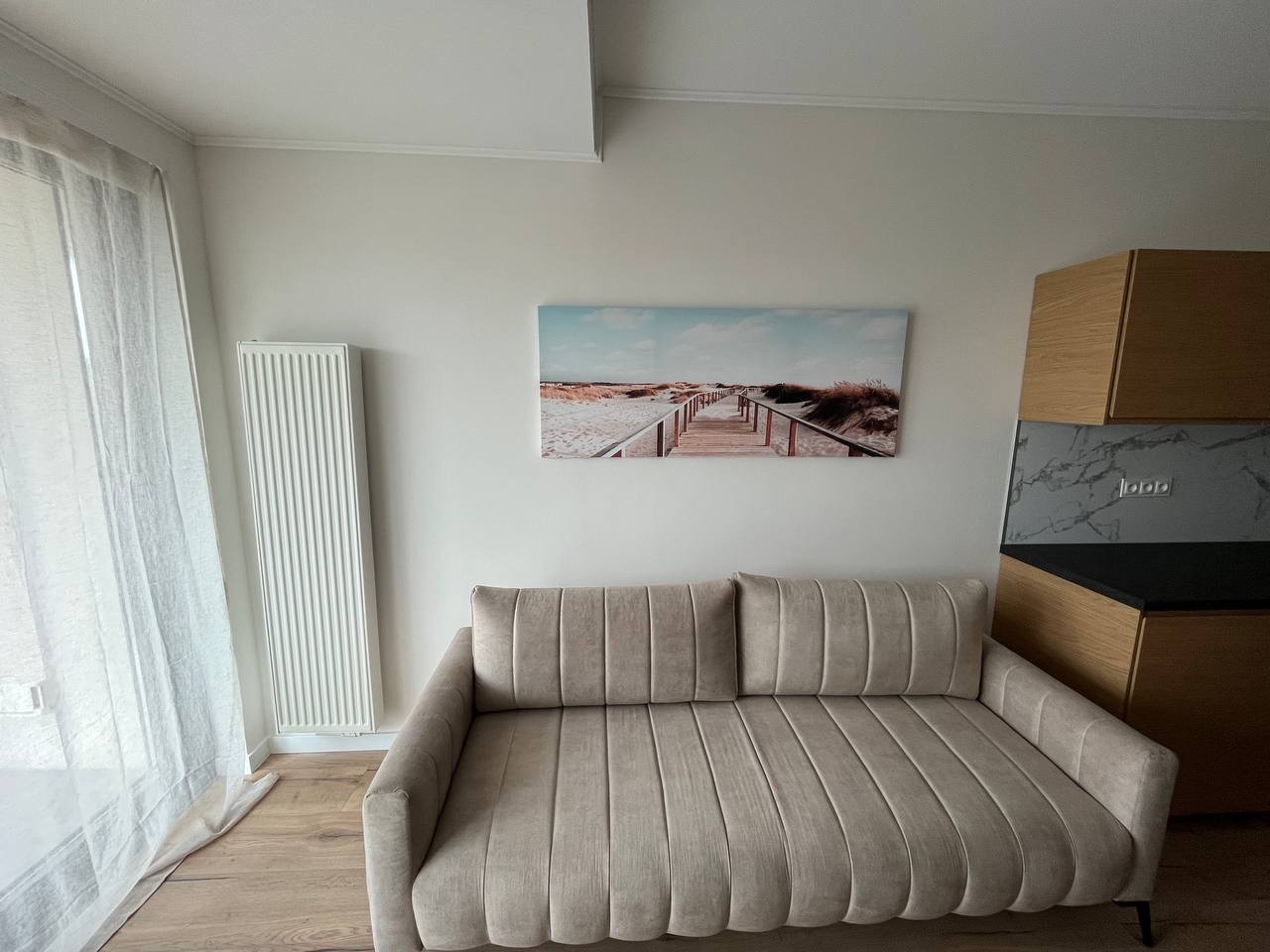 Jasne wnętrze z beżową sofą pod obrazem plaży. Biały grzejnik i zasłony po lewej, drewniane szafki kuchenne po prawej. Minimalistyczny design.