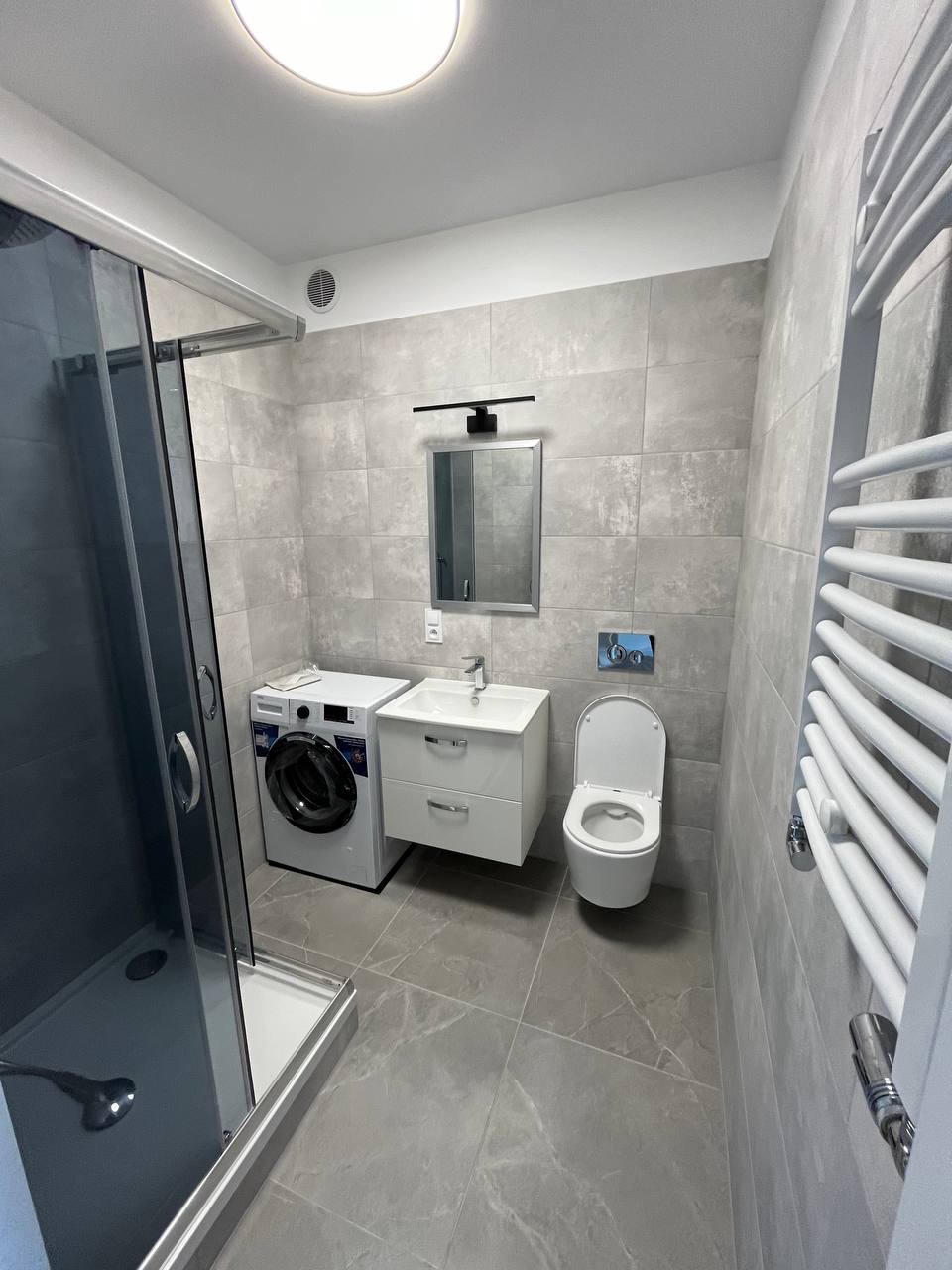 Nowoczesna łazienka w szarościach: kabina prysznicowa z ciemnymi szybami, pralka, umywalka z szafką, toaleta i grzejnik drabinkowy. Minimalistyczny design, płytki imitujące marmur.