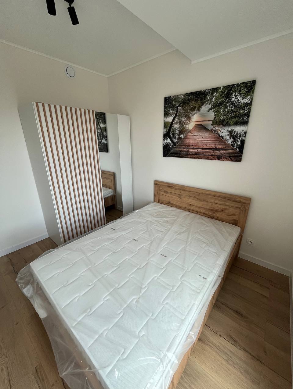 Jasne wnętrze sypialni z drewnianym łóżkiem z materacem w folii, szafą z lamelami i obrazem z motywem molo na ścianie. Podłoga z paneli. Minimalistyczny design.