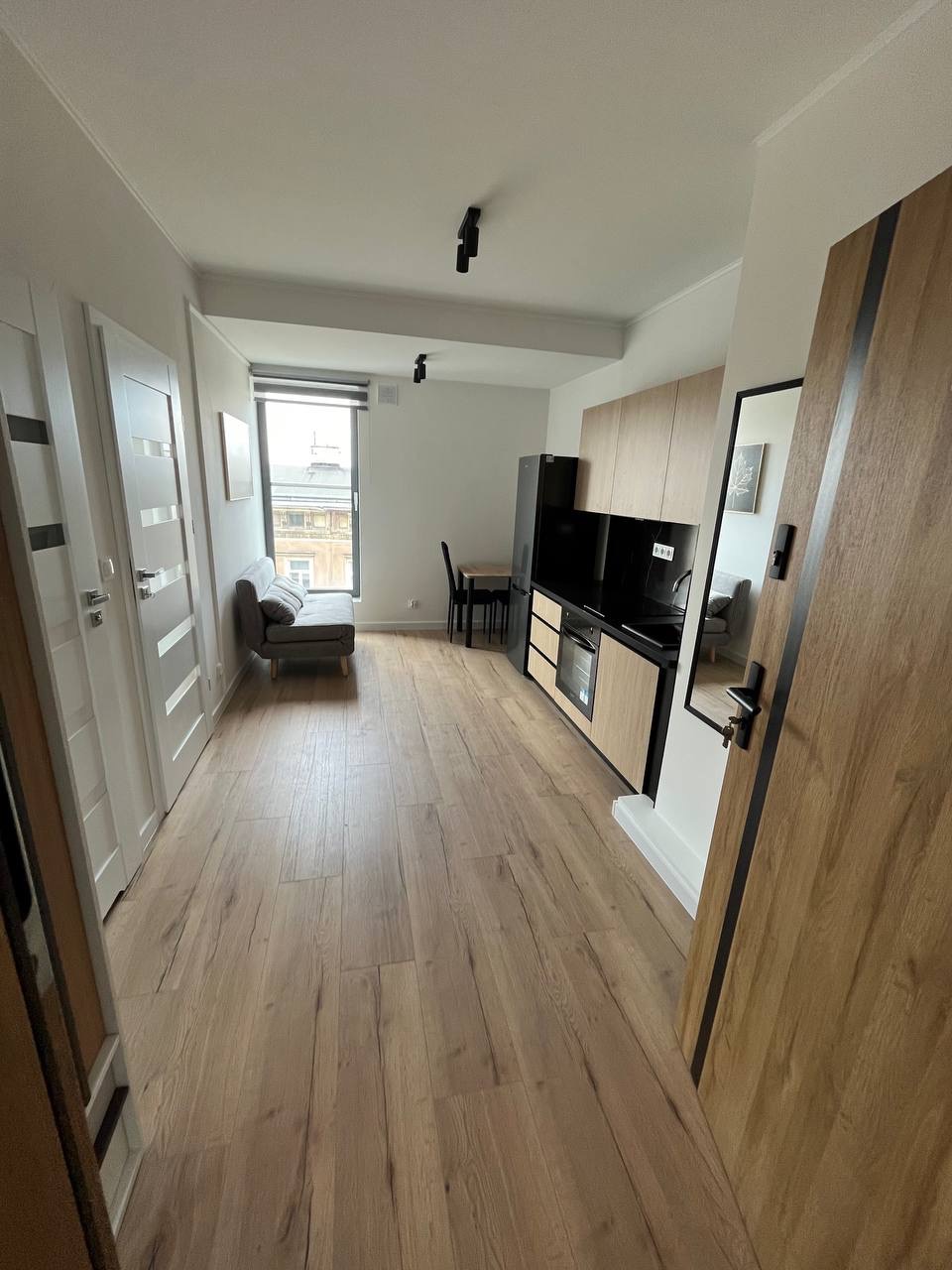 Jasne, nowoczesne wnętrze mieszkania po remoncie: drewniana podłoga, zabudowa kuchenna z czarnym blatem, szara sofa przy oknie. Widok zza otwartych drzwi.