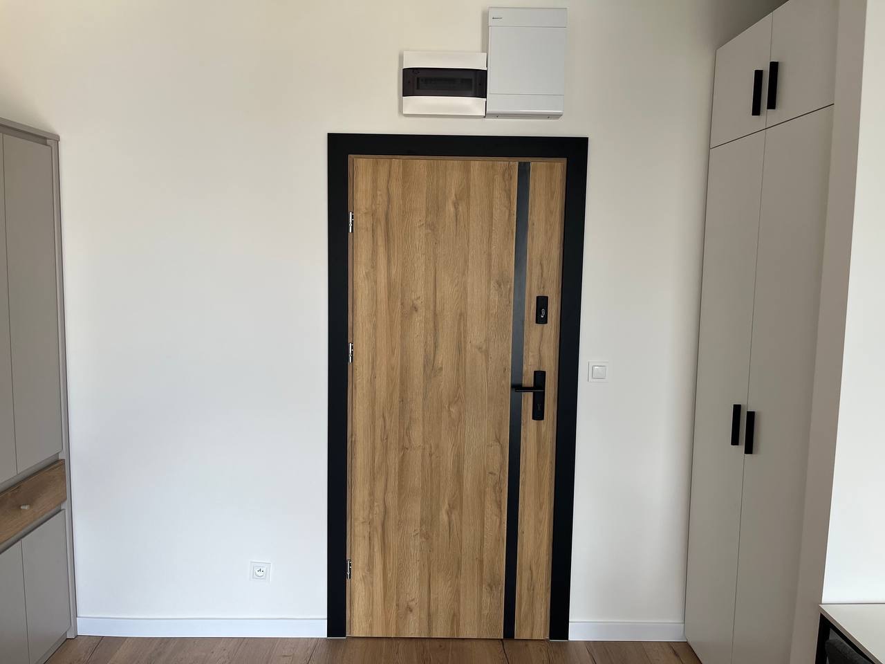 Nowoczesne wnętrze: drewniane drzwi z czarną ramą, białe ściany i szafa w zabudowie. Minimalistyczny design, jasne kolory, widoczna skrzynka elektryczna.