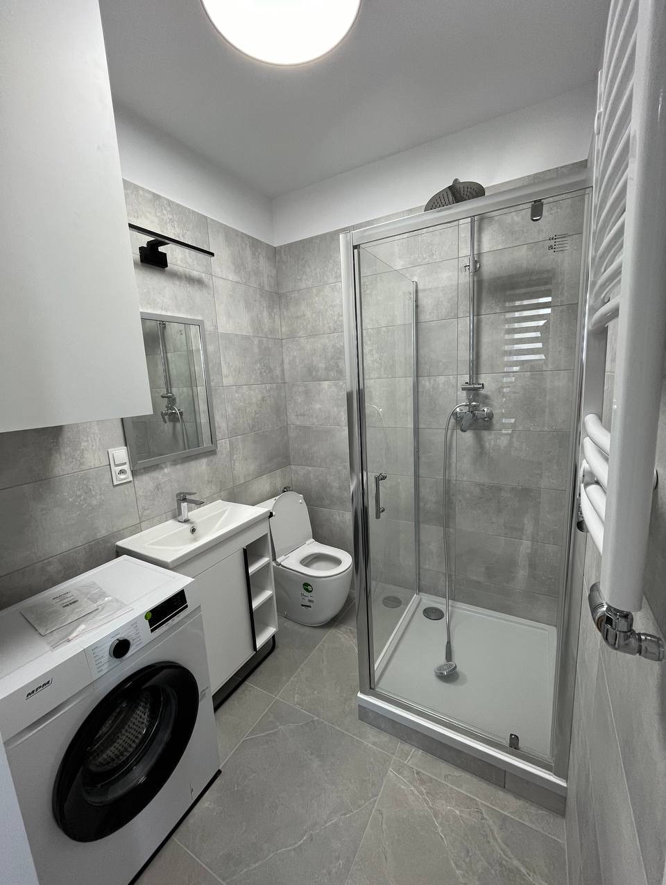 Wykończona łazienka w szarościach: kabina prysznicowa, umywalka z szafką, toaleta i pralka. Minimalistyczny design, płytki imitujące kamień, oświetlenie sufitowe.
