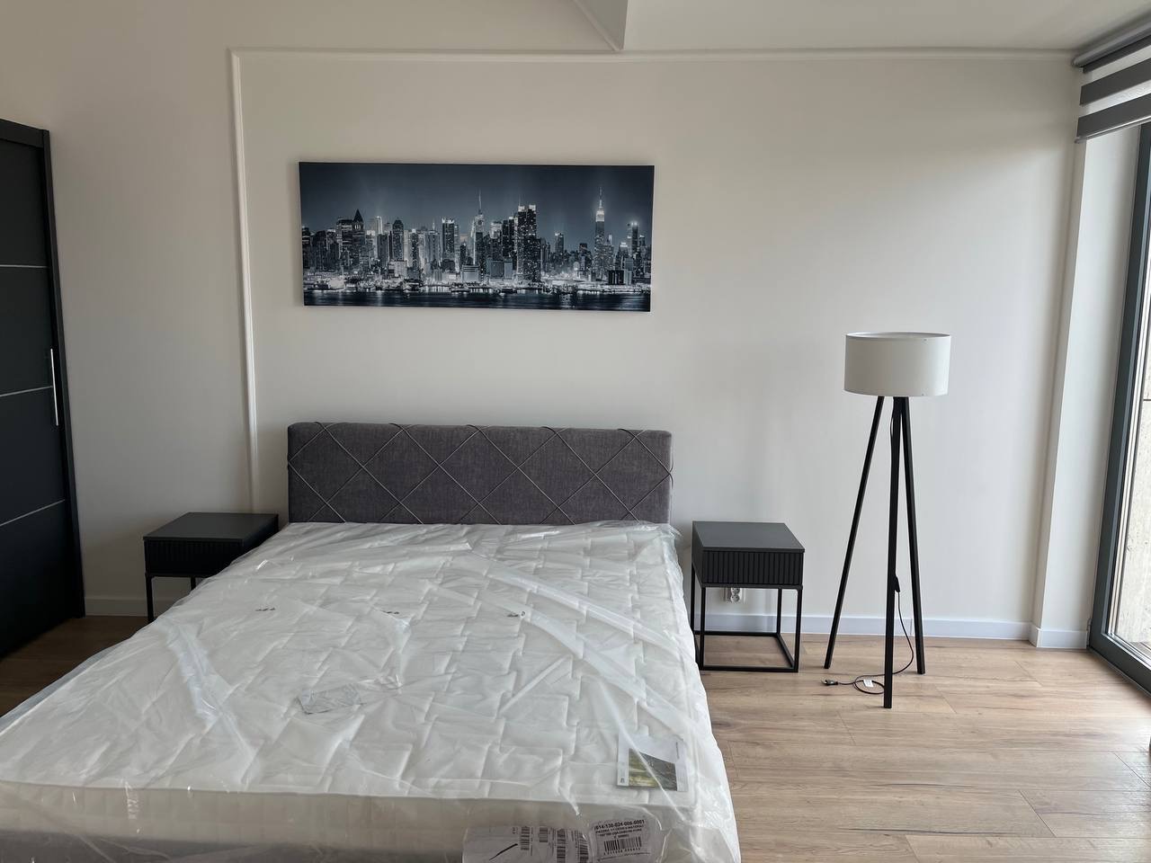 Jasne wnętrze sypialni po remoncie: białe ściany z dekoracyjną listwą, łóżko z szarym zagłówkiem, nocne stoliki i lampa. Na ścianie obraz z panoramą miasta.