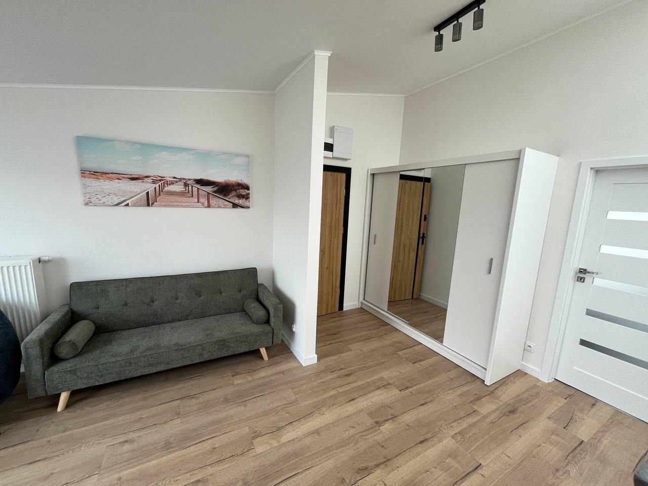 Jasne wnętrze pokoju z drewnianą podłogą, szarą sofą i szafą z lustrzanymi drzwiami. Na ścianie obraz z motywem plaży. Minimalistyczny design.