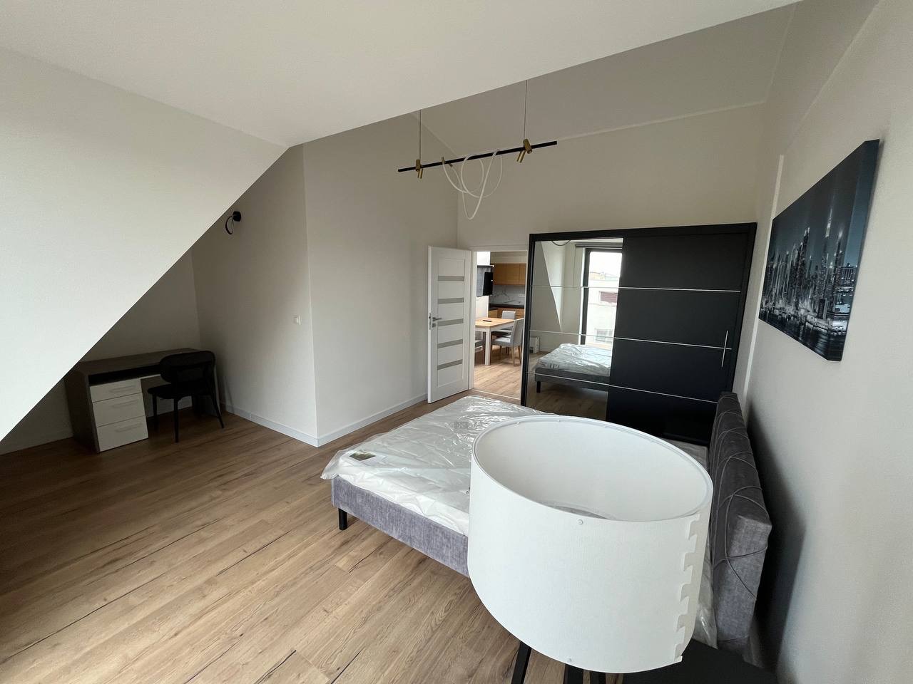 Jasne wnętrze pokoju po odświeżeniu ścian: drewniana podłoga, białe ściany, biurko pod skosem, łóżko z lampą i szafa z lustrem. Widok na kuchnię w tle.