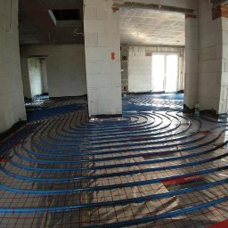 HYDROART Łukasz Zimnoch - Niebieskie rury ułożone spiralnie na folii z czerwoną siatką w surowym wnętrzu budynku w trakcie montażu systemu ogrzewania podłogowego.