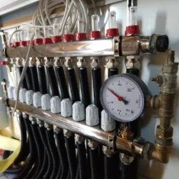 Rozdzielacz ogrzewania podłogowego w szafce, z widocznymi rurami, przepływomierzami z czerwonymi elementami, manometrem i licznymi złączkami.