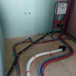 Wnętrze łazienki w trakcie prac hydraulicznych: widoczne rury wodne w kolorach czerwonym i niebieskim, rura kanalizacyjna karbowana z pomarańczowymi uchwytami oraz stelaż podtynkowy TECE.