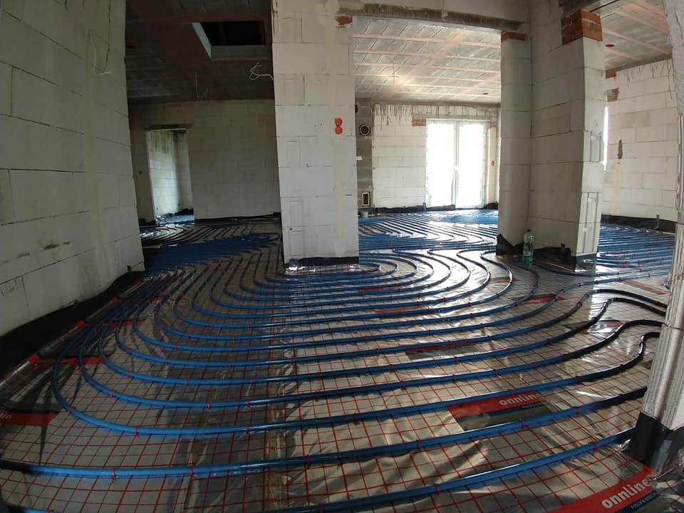 Niebieskie rury ułożone spiralnie na folii z czerwoną siatką w surowym wnętrzu budynku w trakcie montażu systemu ogrzewania podłogowego.