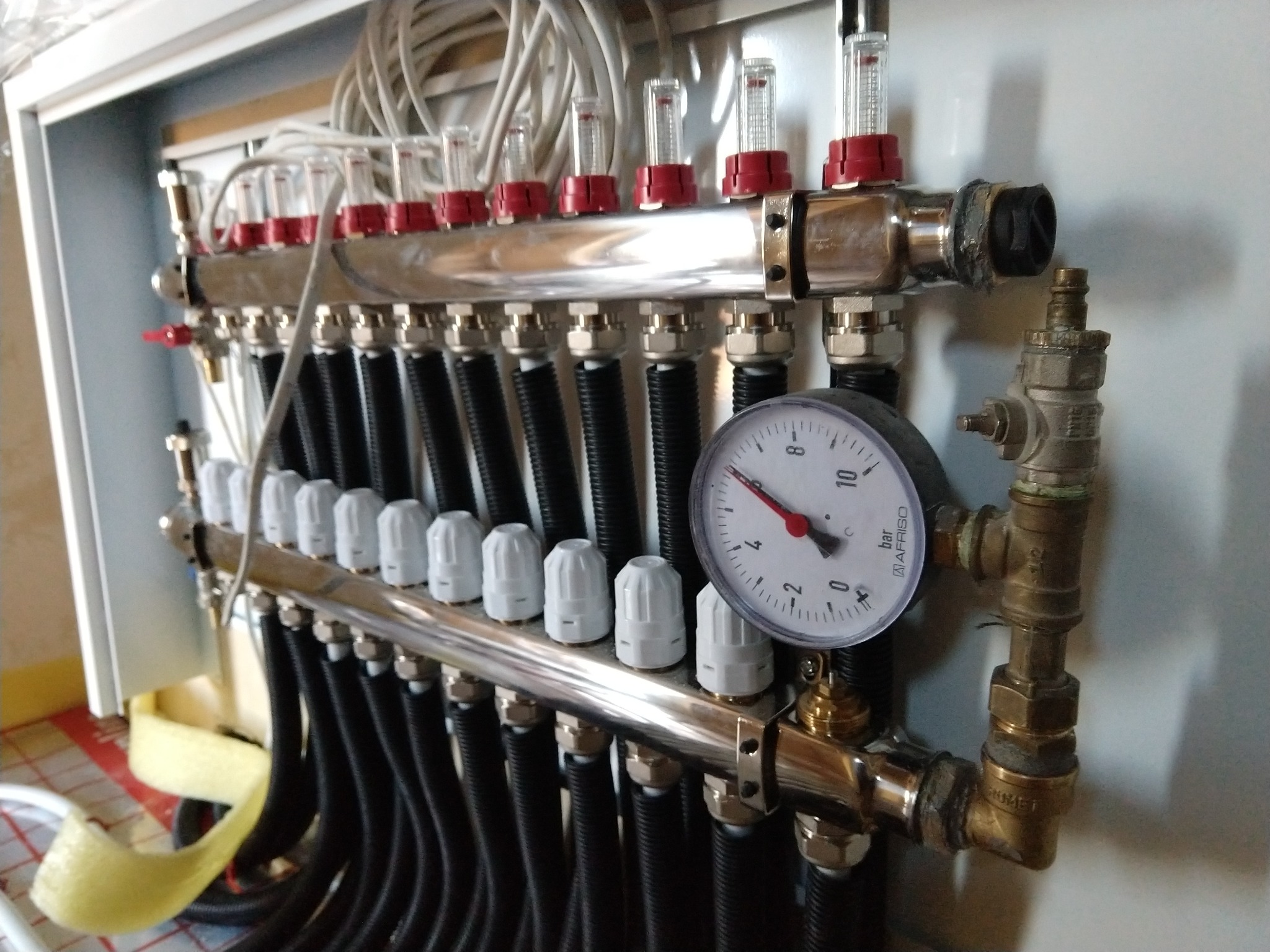 Rozdzielacz ogrzewania podłogowego w szafce, z widocznymi rurami, przepływomierzami z czerwonymi elementami, manometrem i licznymi złączkami.