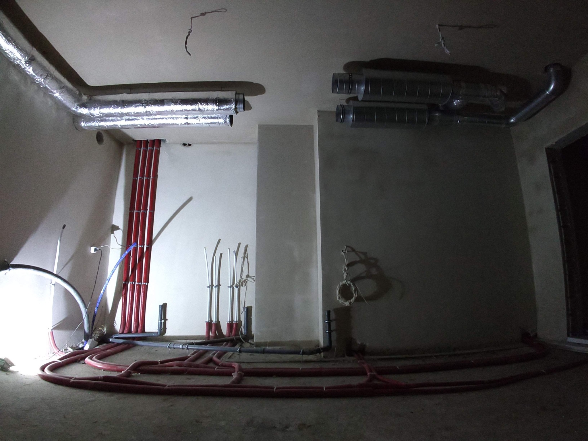Wnętrze w trakcie prac instalacyjnych: widoczne czerwone rury ogrzewania podłogowego na posadzce, pionowe rury grzewcze przy ścianie oraz kanały wentylacyjne pod sufitem, częściowo zaizolowane...