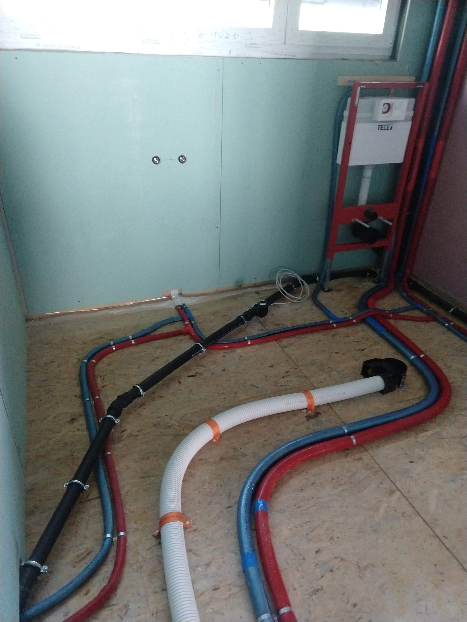 Wnętrze łazienki w trakcie prac hydraulicznych: widoczne rury wodne w kolorach czerwonym i niebieskim, rura kanalizacyjna karbowana z pomarańczowymi uchwytami oraz stelaż podtynkowy TECE.