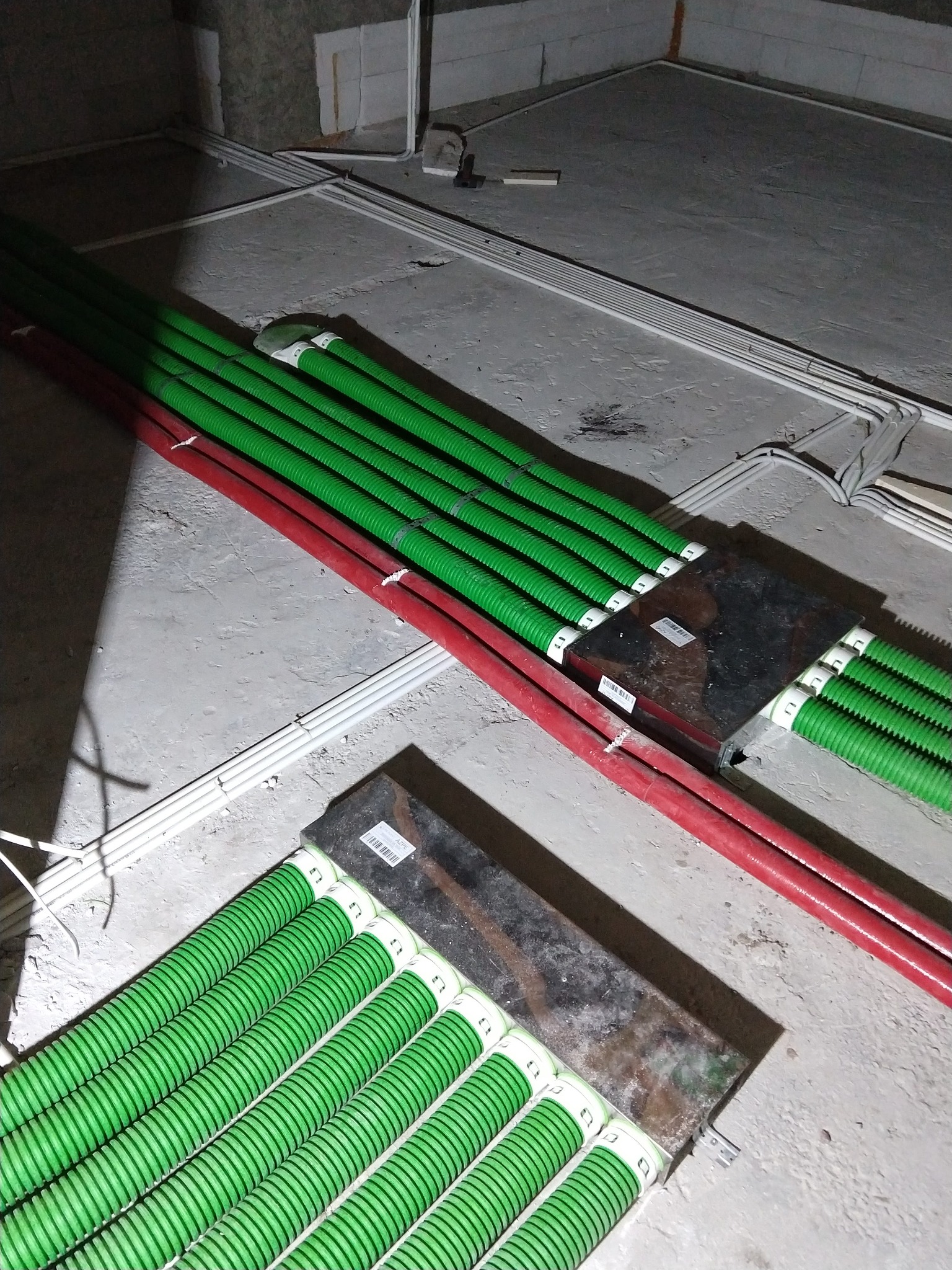 Wiązki zielonych i czerwonych rur karbowanych, układane na surowym, betonowym podłożu w trakcie prac instalacyjnych, widoczne metalowe skrzynki łączeniowe.