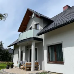 Nowoczesny dom z balkonem zabezpieczonym szkłem mlecznym i metalową poręczą, widoczne meble ogrodowe na tarasie z kostki brukowej.