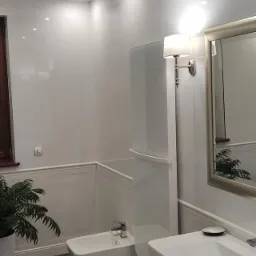 Nowoczesna łazienka z białymi płytkami, ciemną marmurową podłogą, białym bidetem i umywalką z szafką, oświetlona punktowymi lampami sufitowymi i kinkietem z abażurem, dekorowana zieloną rośliną...