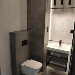 Nowoczesna łazienka z podwieszaną toaletą, umywalką na szafce z otwartymi półkami i szarą płytką imitującą kamień na ścianach.