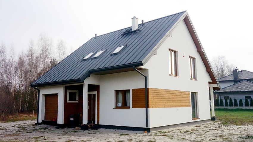 Nowoczesny dom jednorodzinny z białymi ścianami, grafitowym dachem z trzema oknami dachowymi i elementami drewnianej elewacji, widoczny garaż z brązową bramą.