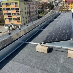 Panele fotowoltaiczne zamontowane na płaskim dachu budynku mieszkalnego, widok z góry na instalację, z betonowymi bloczkami i przewodami w peszlu, w tle zabudowa miejska.