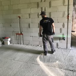 El-mar instalacje elektryczne - Elektryk w trakcie pracy w surowym wnętrzu, widoczne pionowe rury instalacyjne w ścianie z betonu komórkowego oraz laserowy niwelator wyznaczający poziom.