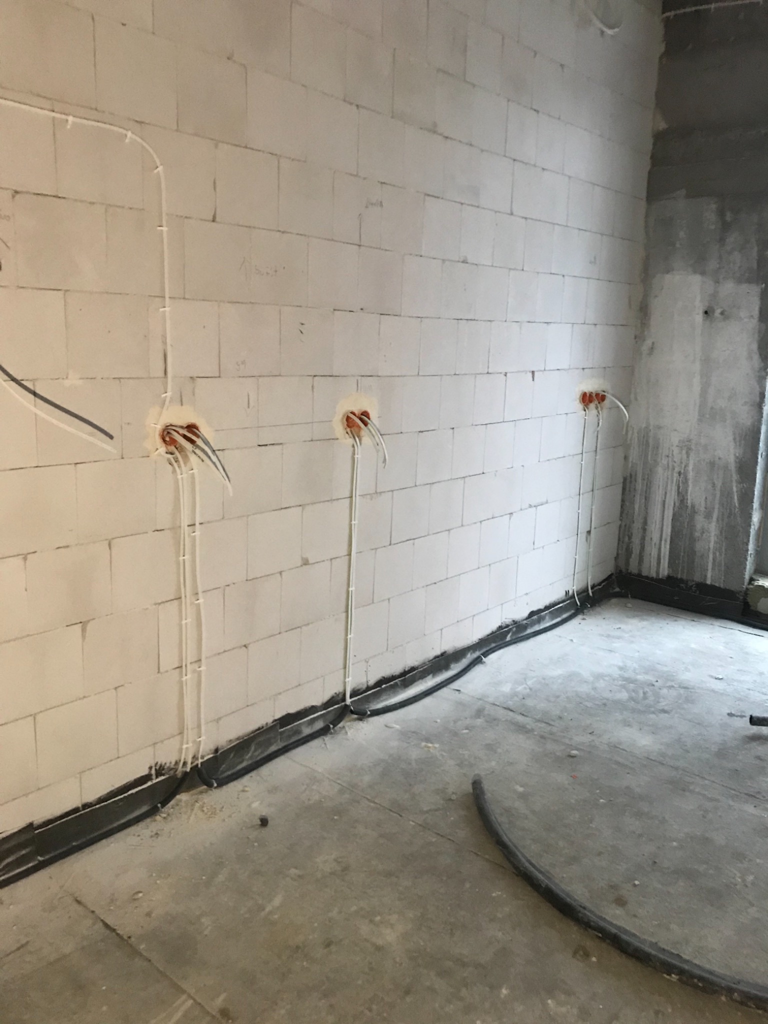Surowy stan wnętrza z widocznymi przewodami elektrycznymi wychodzącymi ze ścian z bloków betonowych, zabezpieczonymi pianką montażową, oraz kablami ułożonymi wzdłuż ściany i na podłodze.