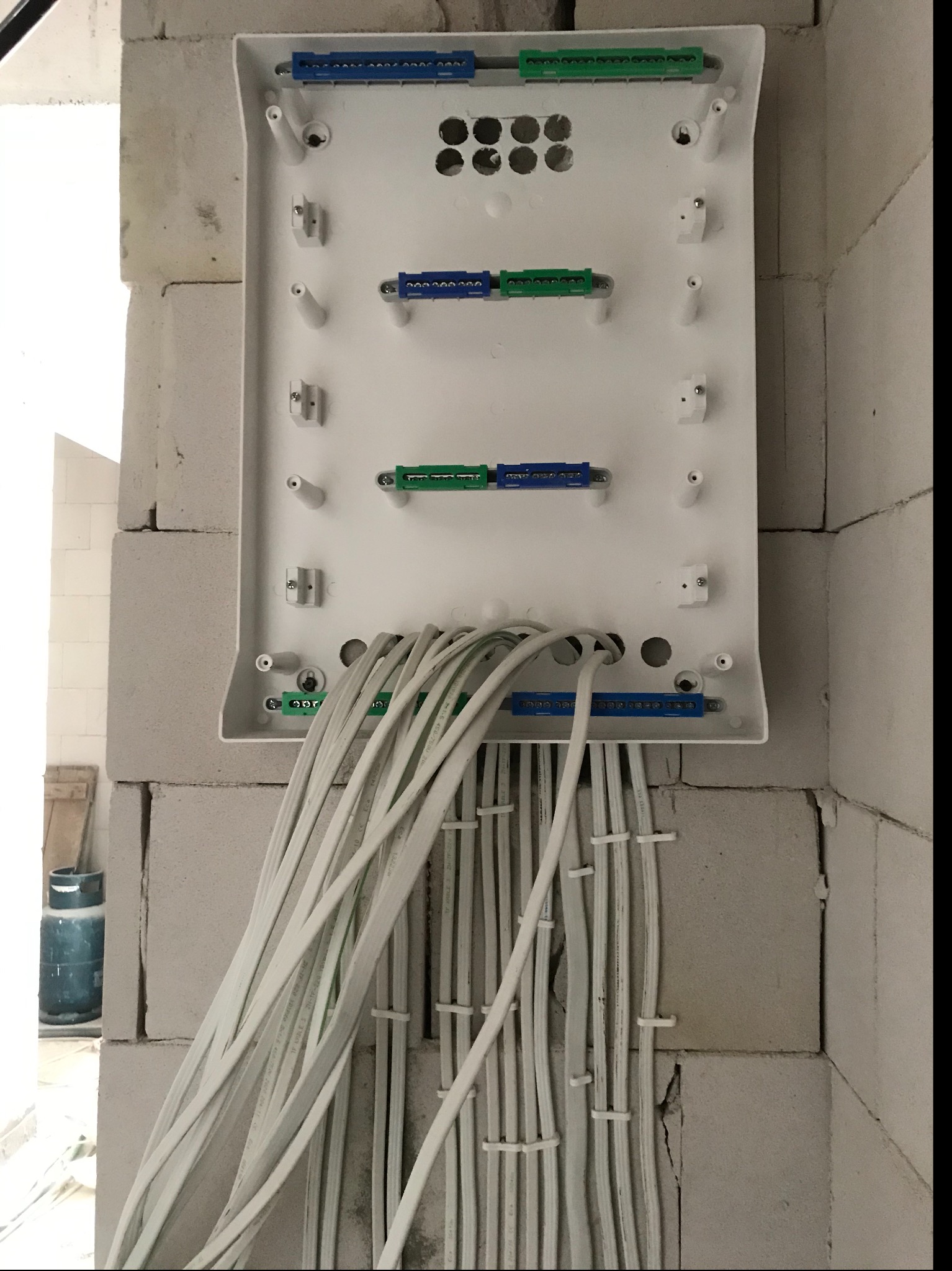 Biała skrzynka rozdzielcza zamontowana na ścianie z białych bloczków, z licznymi białymi kablami wchodzącymi do niej od dołu. Kable przymocowane do ściany za pomocą plastikowych uchwytów. Widoczne...