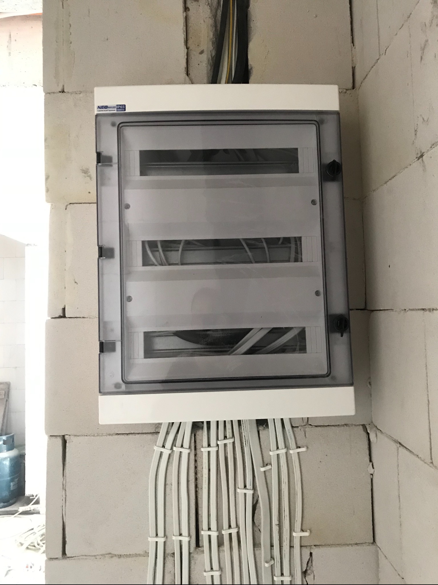 Biała skrzynka elektryczna z przezroczystą pokrywą zamontowana na ścianie z białych bloczków, z widocznymi kablami wewnątrz i wychodzącymi na zewnątrz w peszlach, w surowym stanie wykończenia.