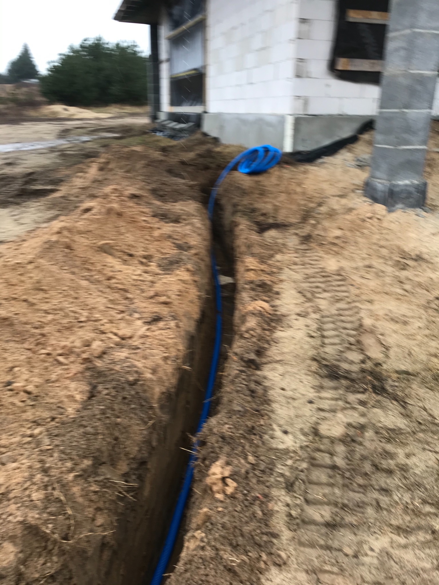 Niebieski przewód w wykopie ziemnym, w tle budynek w trakcie budowy z betonowych bloków.