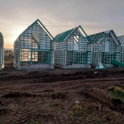 Wczesny etap budowy kilku domów szkieletowych z zielonymi elementami konstrukcyjnymi i białymi panelami, widoczne ślady opon w błotnistym terenie, maszyna budowlana w oddali pod pochmurnym niebem.