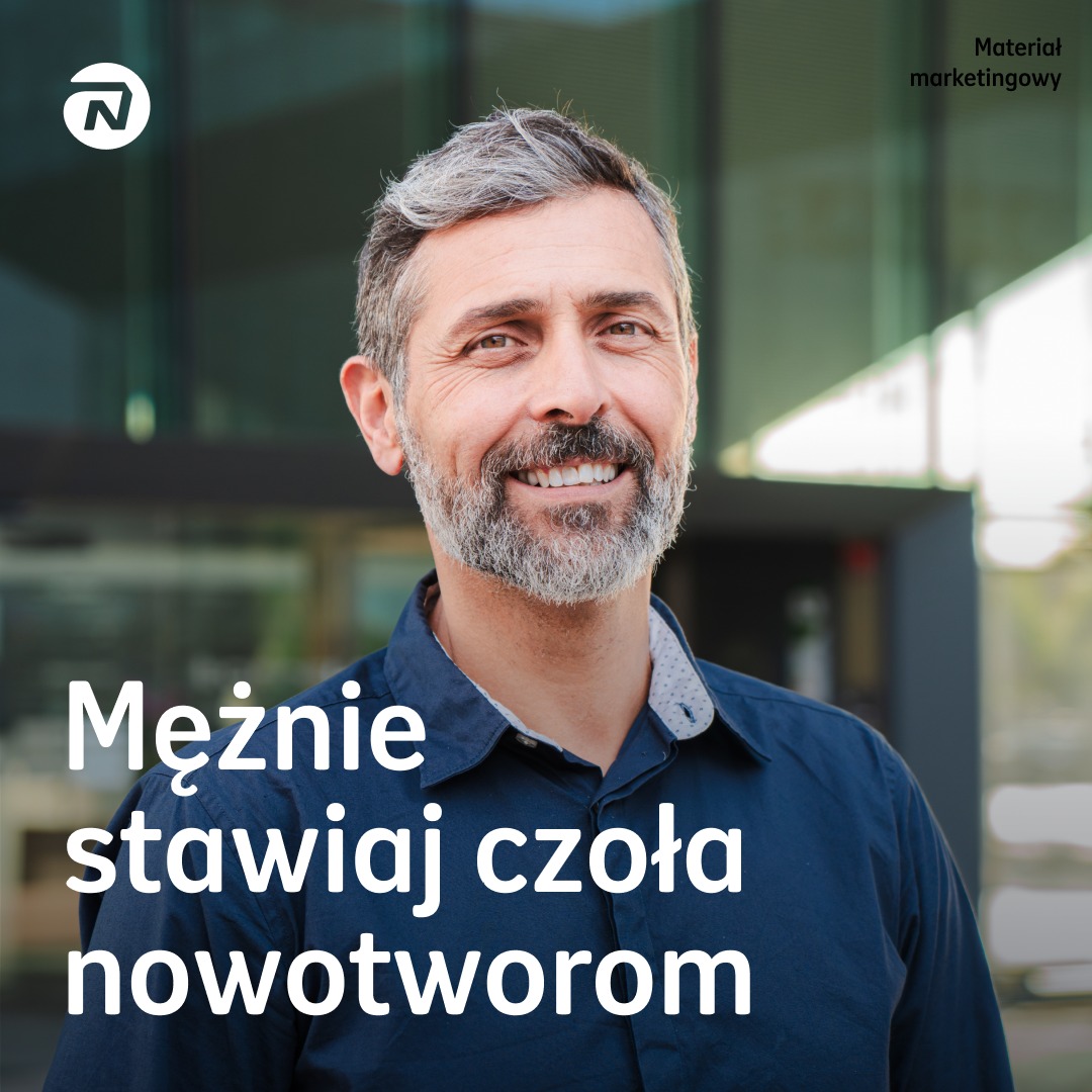 Uśmiechnięty mężczyzna w średnim wieku z siwiejącą brodą i włosami, ubrany w ciemną koszulę, na tle nowoczesnego budynku. Materiał marketingowy.